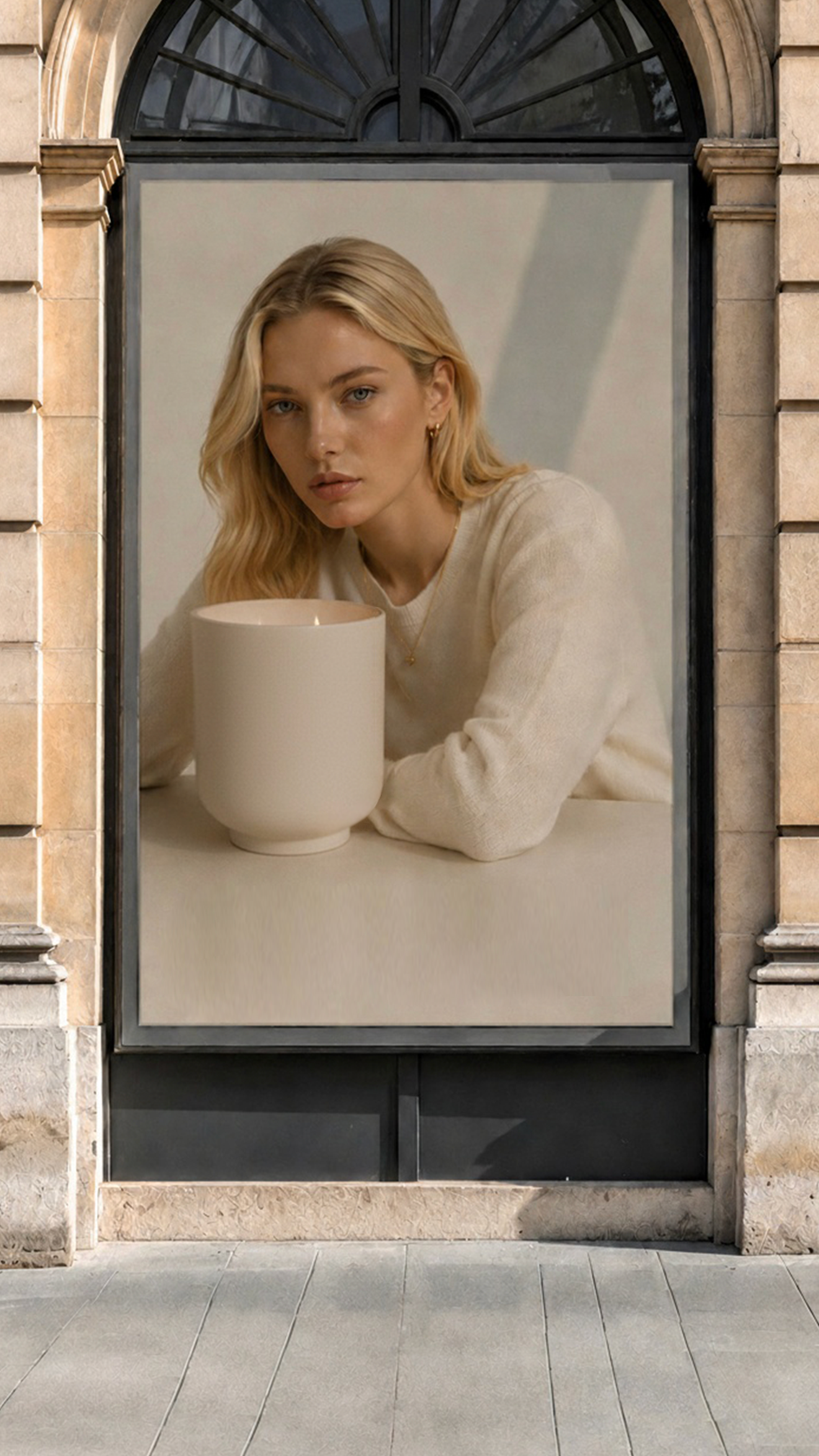 A large vertical wall poster or art print mounted on a stone building facade, featuring a blonde woman in a cream sweater leaning over a table with a large cream ceramic rene refillable vase.